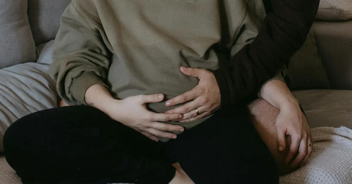 IVF clinic for couples with a pregnant person sitting on a couch, embraced by a partner with hands on their belly.