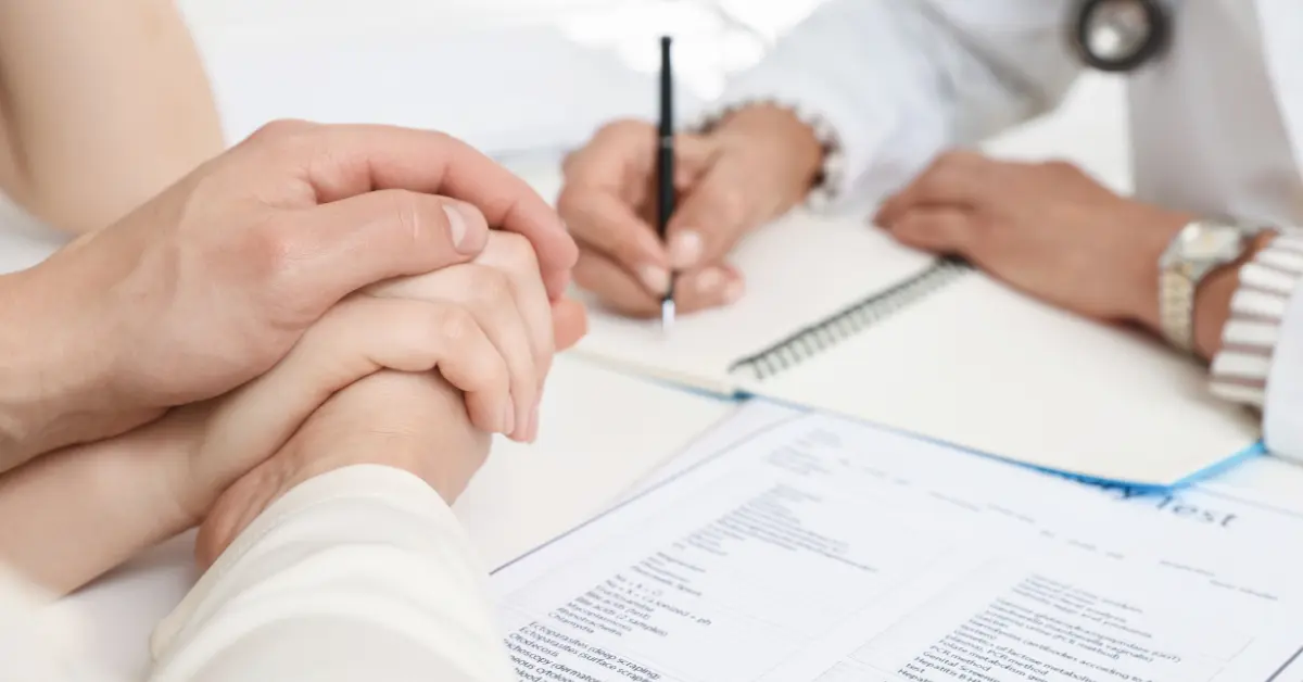 Fertility and age in women discussed with a doctor while a couple holds hands across a desk with medical forms.