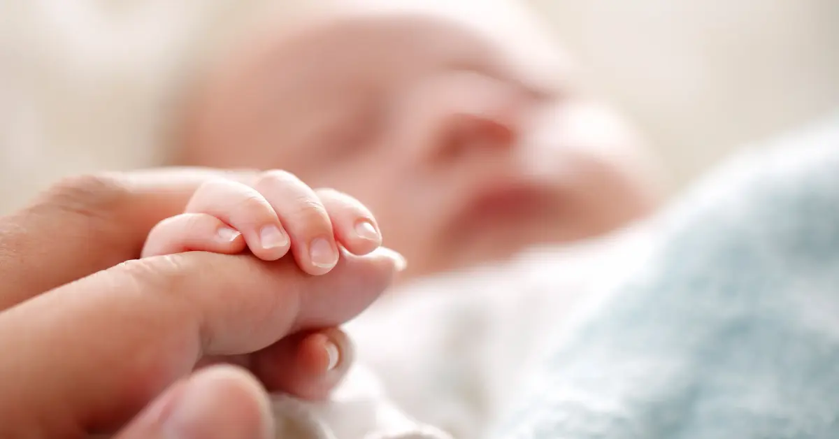 Newborn baby holding an adult's finger while resting peacefully in a soft blanket